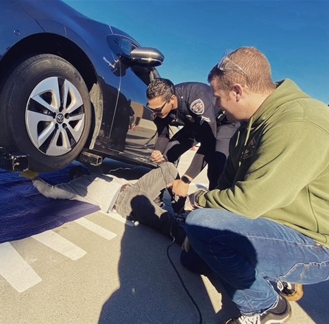 two police officers etching a serial number on to a catalytic converter under a vehicle with a person watching next to the vehicle